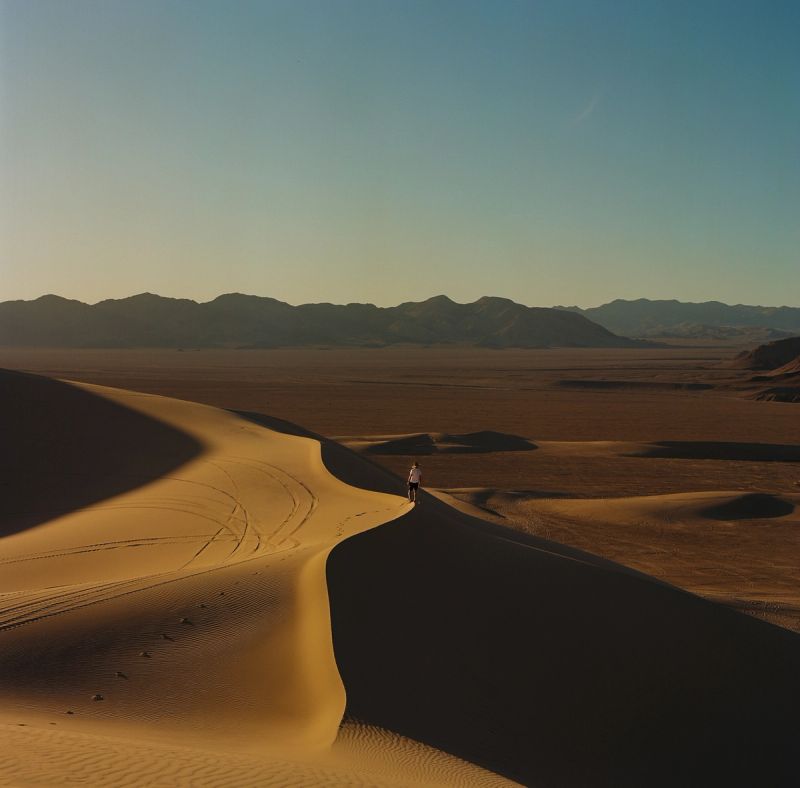 A lone figure walks along the crest of a large sand dune in a vast desert landscape.