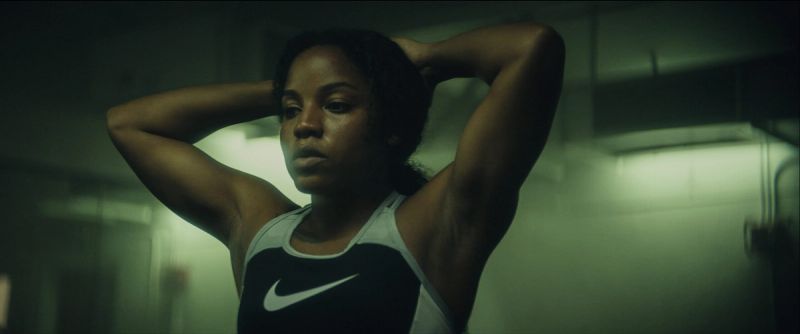 Female athlete Jen Welter poses with hands behind head wearing a black Nike tank top in a dimly lit room.