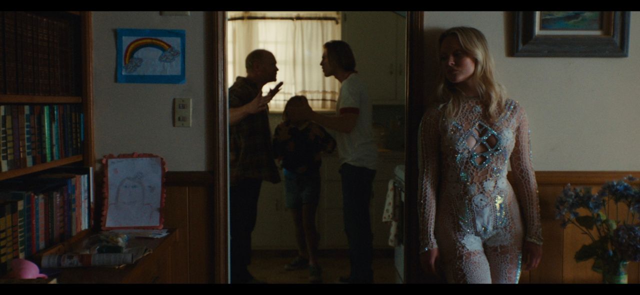 Zara Larsson in a shimmering lace bodysuit standing in a dimly lit home with family arguing in the background.
