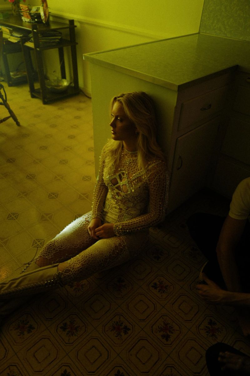 Zara Larsson in a shimmering gold bodysuit sits on a patterned tile floor, leaning against a kitchen counter under warm yellow lighting.