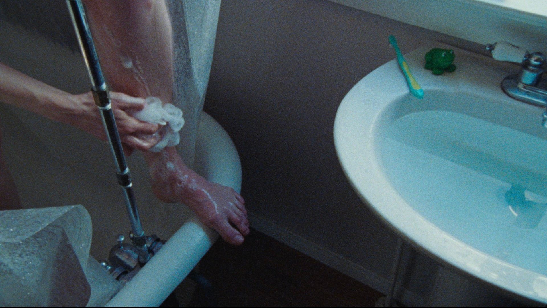 Close-up of bare feet being washed with soap in a bathtub, sink and toothbrush visible nearby.