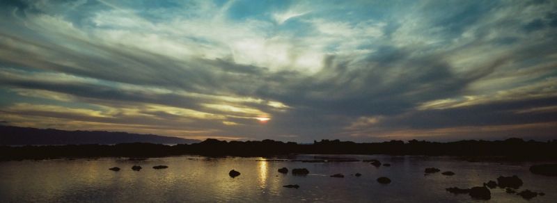 Sunset over calm water with distant hills and scattered rocks