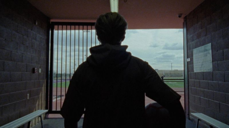 Silhouette of a person in hoodie looking out at an outdoor track and field from a brick corridor