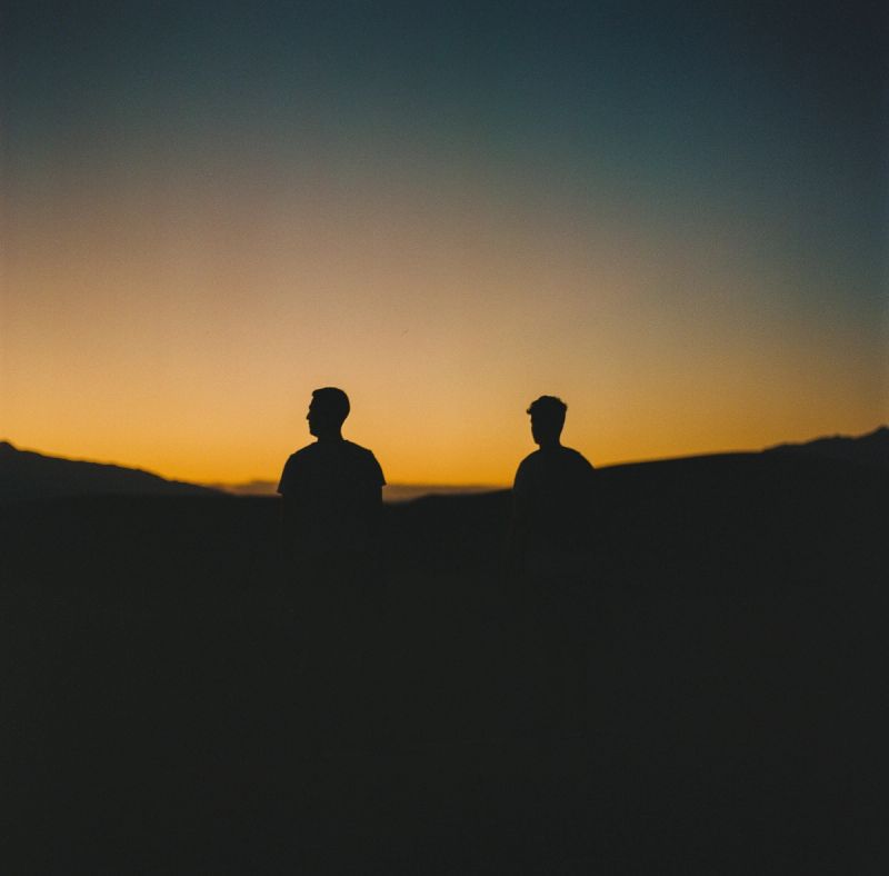 Two men silhouetted against a golden sunset horizon in a mountain landscape.