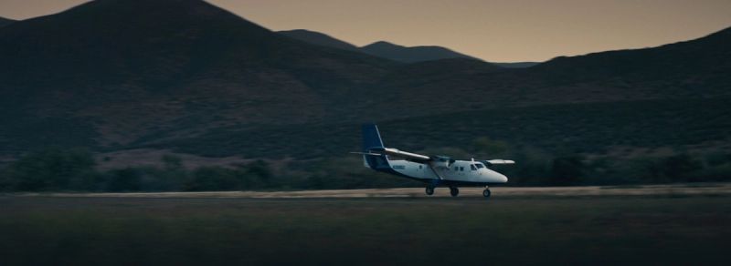 A white twin-engine propeller plane taxis on a runway with mountains in the background.