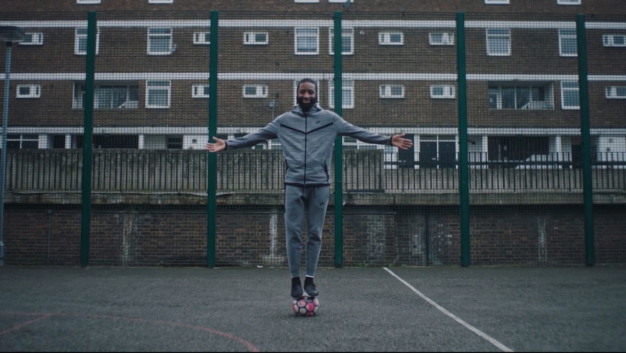 Man in grey Nike tracksuit balancing on soccer ball with arms outstretched on outdoor court