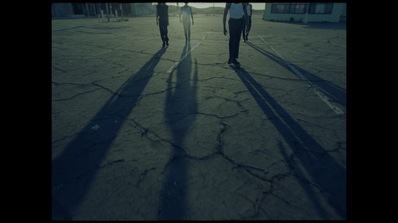 Four figures walking across cracked asphalt with long shadows at low light