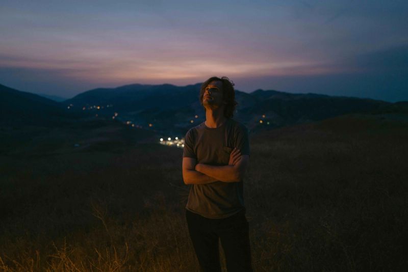 Man stands with arms crossed on hillside at twilight overlooking distant town lights