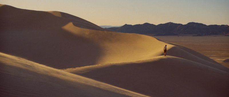 A lone figure walks across golden sand dunes at sunset in the desert.
