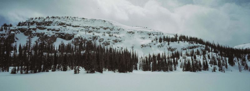 Snow-covered mountain ridge with dark evergreen trees under cloudy skies.