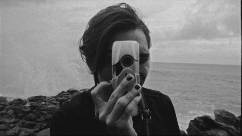 A person holding a compact camera against a rocky shoreline backdrop.
