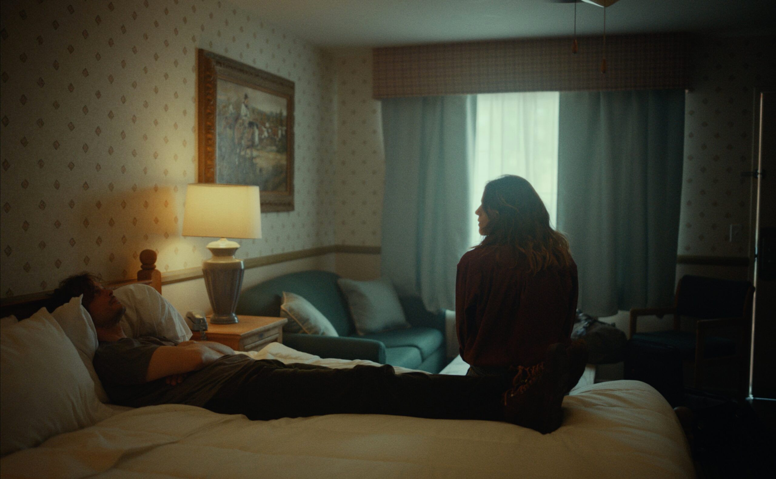 A couple in a dimly lit hotel room, man lying on bed, woman sitting by window.