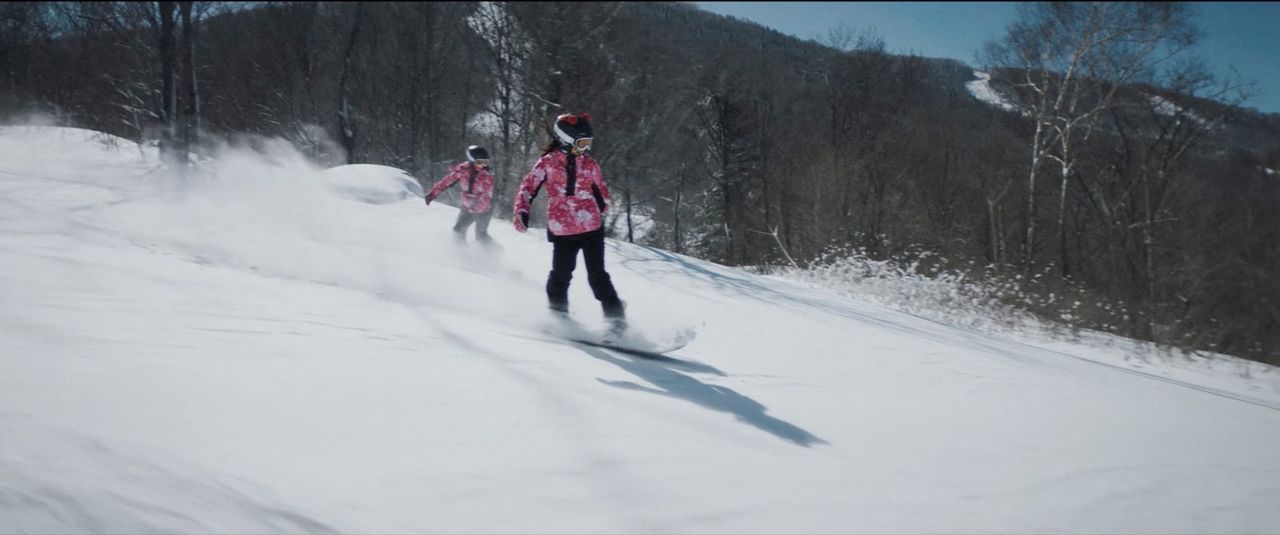Two snowboarders in pink jackets carve down a snowy slope with trees and mountains in the background.
