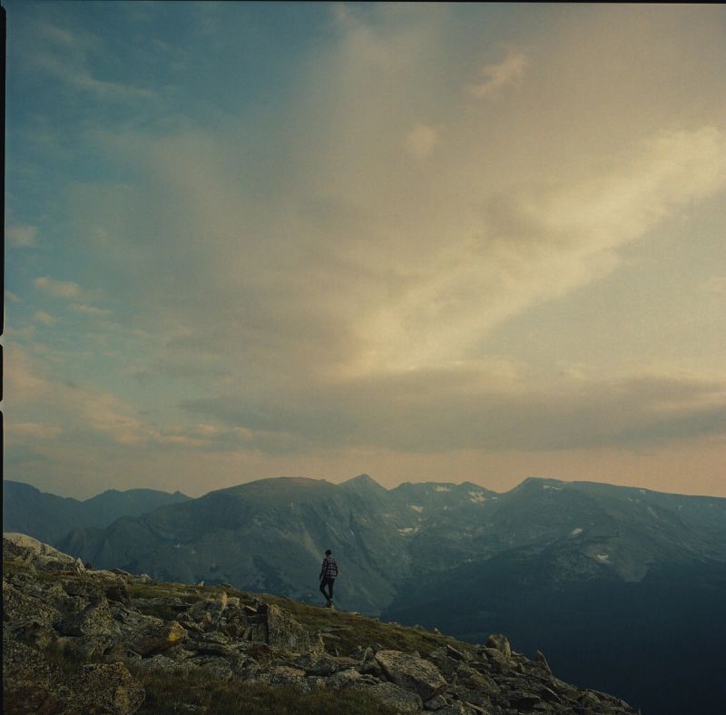 Solitary figure stands on rocky mountain ridge overlooking vast peaks under cloudy sky, shot on film.