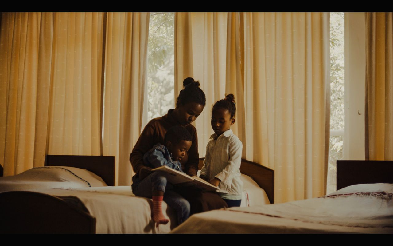 Mother reading a book to two children in a warmly lit hotel room with yellow curtains.