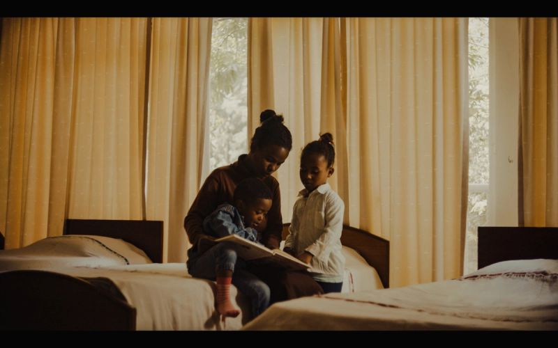 Mother reading a book to two children in a warmly lit hotel room with yellow curtains.