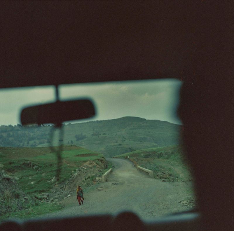 View through car windshield of lone figure walking on rural dirt road in green hills.