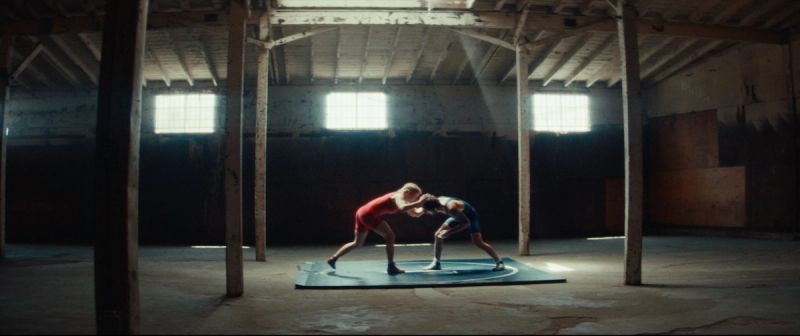 Two female wrestlers grappling on a blue mat inside a rustic warehouse with bright window light.