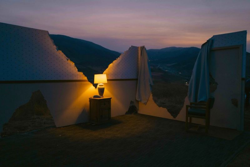 Warm lamp glows beside a torn wall overlooking mountain valleys at dusk