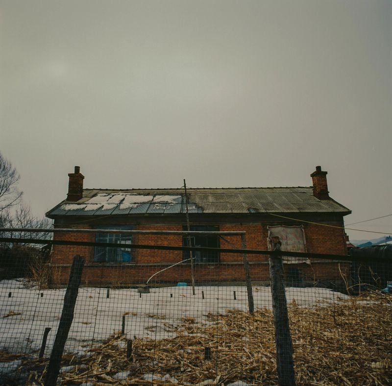 A weathered brick house with snow on the roof stands behind a wire fence in a snowy landscape.