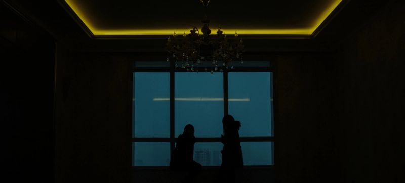 Silhouettes of two people standing by a large window in a dimly lit room with yellow ceiling lights.