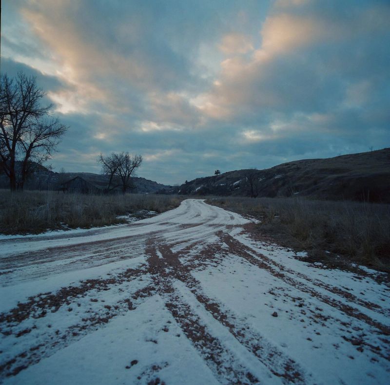 Snow-dusted dirt road winding through hilly landscape under cloudy sky