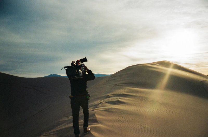 Cinematographer with camera gear walking across sunlit desert dunes