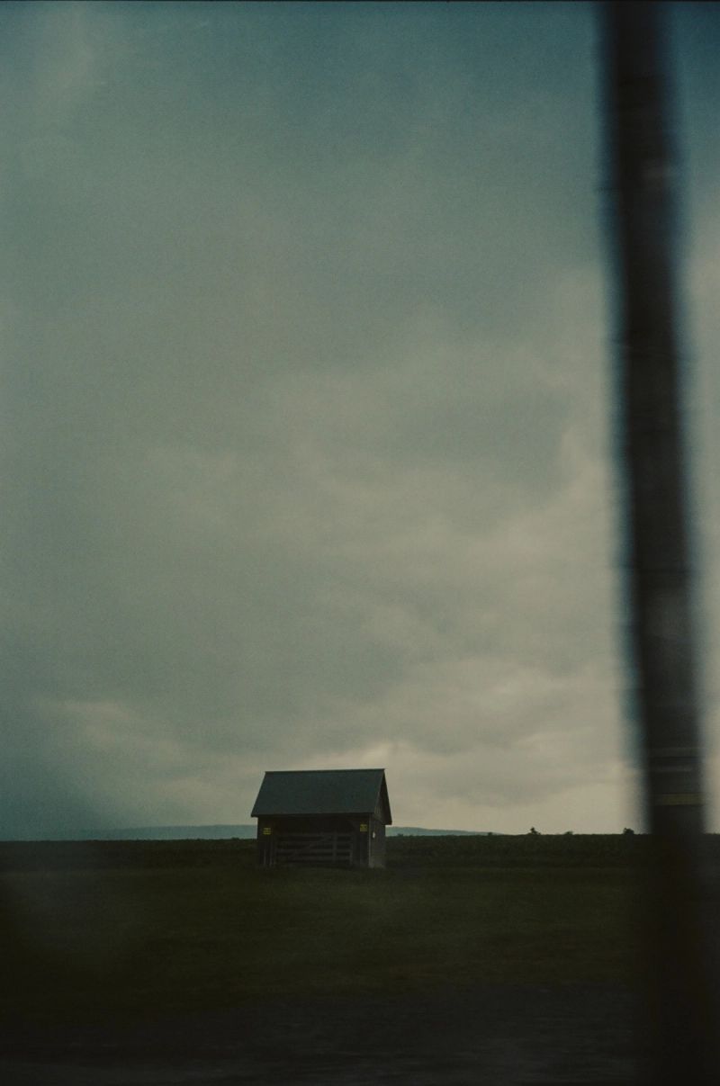 A solitary wooden shed stands in a flat green field under an overcast sky.