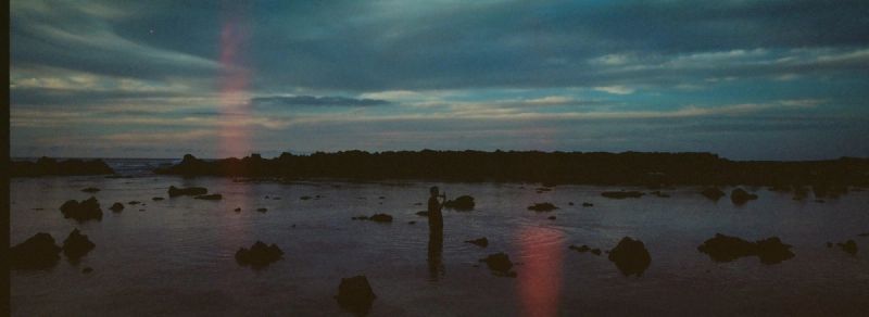 Cinematic silhouette of a figure standing in shallow water on a rocky beach at twilight.
