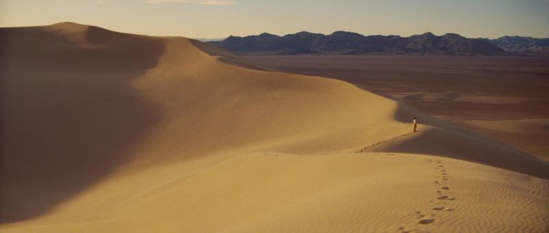 A lone figure walks across golden desert dunes with distant mountains under a soft sky.