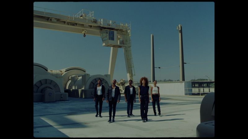Five people walking toward camera in industrial port with cranes and large machinery under blue sky