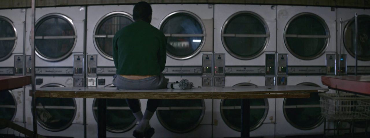 Person sitting on table in dimly lit laundromat with rows of washing machines.