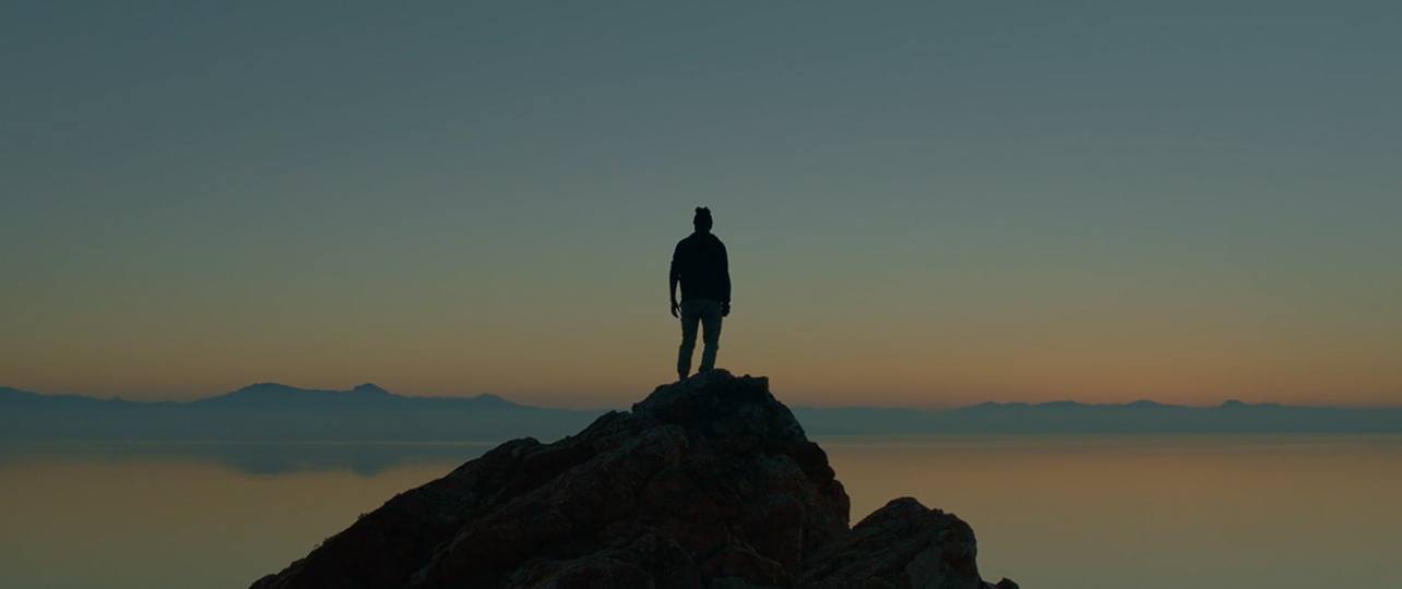 Silhouette of a person standing on a rocky peak at dusk over calm water and distant mountains