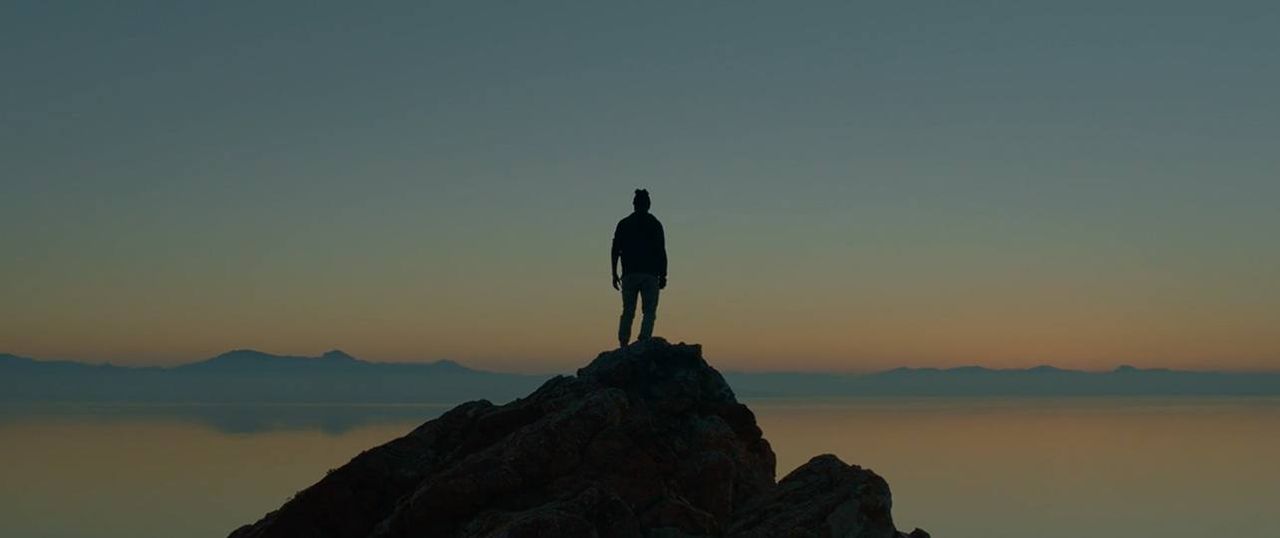 Silhouette of a person standing on a rocky peak at dusk over calm water and distant mountains