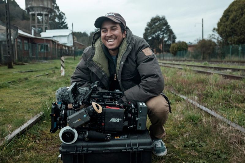 Cinematographer smiles while crouching next to a large ARRI Alexa camera setup on train tracks.