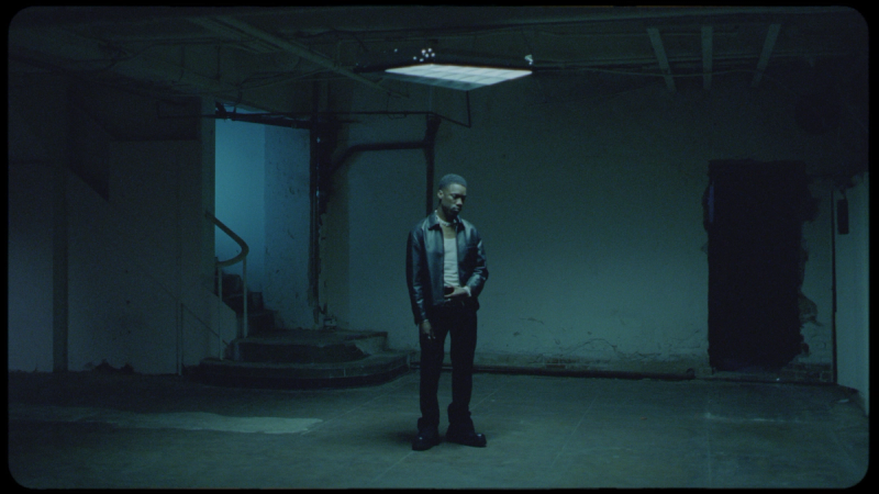 Giveon stands alone in a dimly lit, industrial-style basement with blue-tinted lighting.