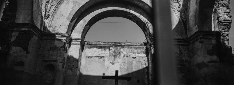 Black and white film photography of a weathered stone archway with a cross, captured in Guatemala.