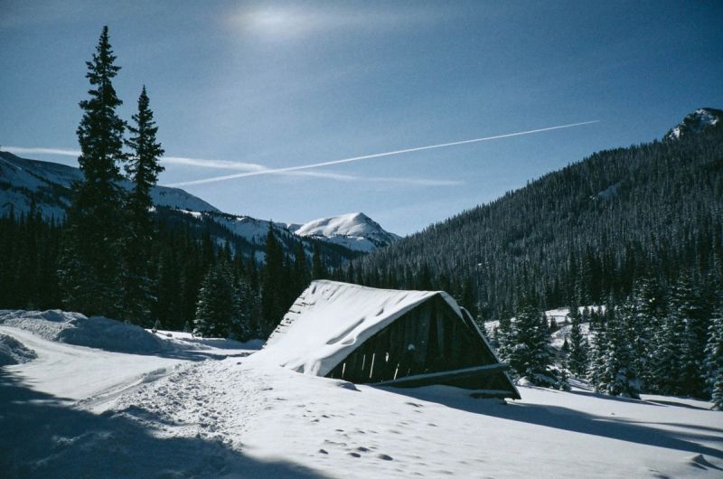 Snow-covered wooden cabin nestled among pine trees in a winter mountain landscape under blue sky.