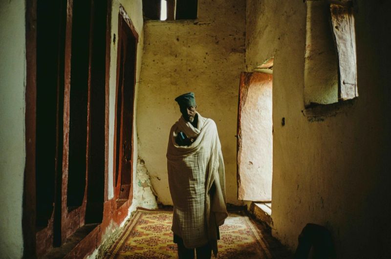 Elderly man wrapped in a shawl stands in a dim, rustic interior hallway with natural light.