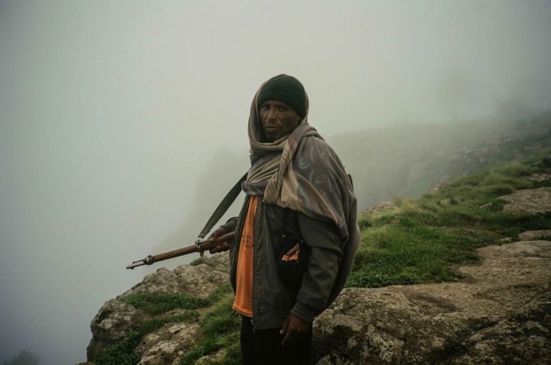 Film photography of a man holding a rifle standing on a foggy, rocky mountain ridge.