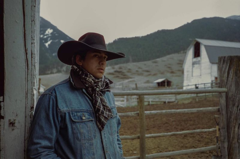 Young man in cowboy hat and denim jacket stands by wooden fence with mountains and barn in background