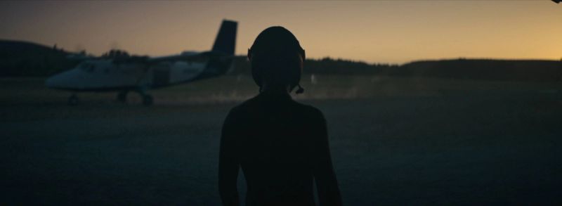 Silhouette of a skydiver in helmet facing a small propeller plane on an airfield at dusk.