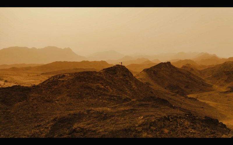 Cinematographer stands atop rugged mountain peak in golden desert landscape