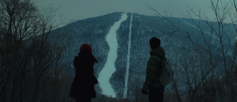 Two figures stand before a snow-covered mountain with ski runs under overcast skies.