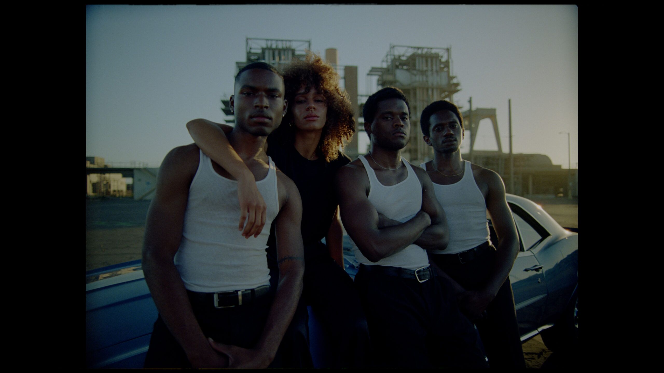 Four Black models in white tank tops posing by a vintage car at industrial sunset