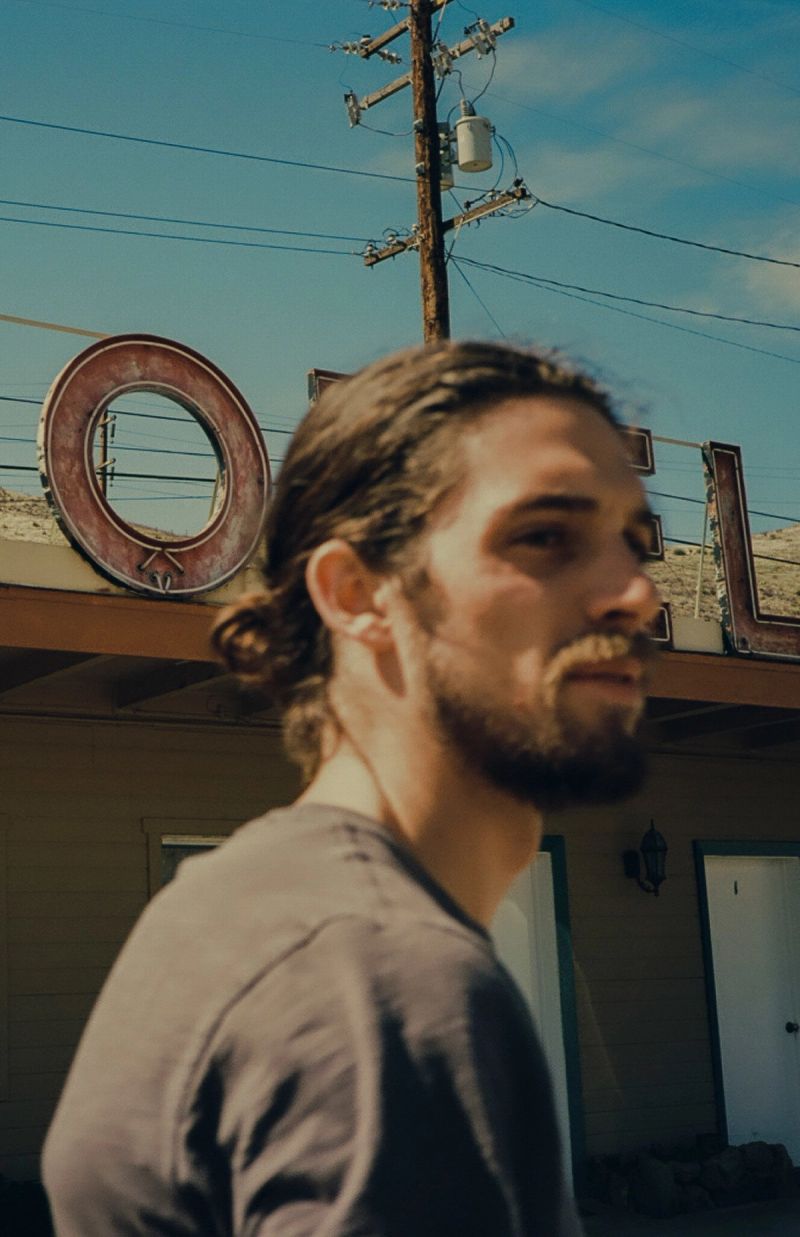 Man with beard and ponytail in front of rustic building under blue sky