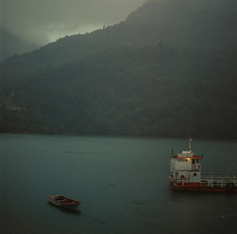 A misty lake scene with a large boat and small rowboat near forested mountains.