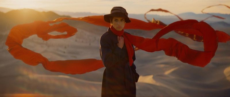 Woman in hat and coat with flowing red scarf against desert dunes at sunset