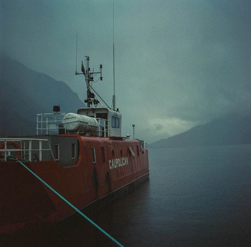 Red workboat named Caupolican moored in a misty Patagonian fjord with mountains.