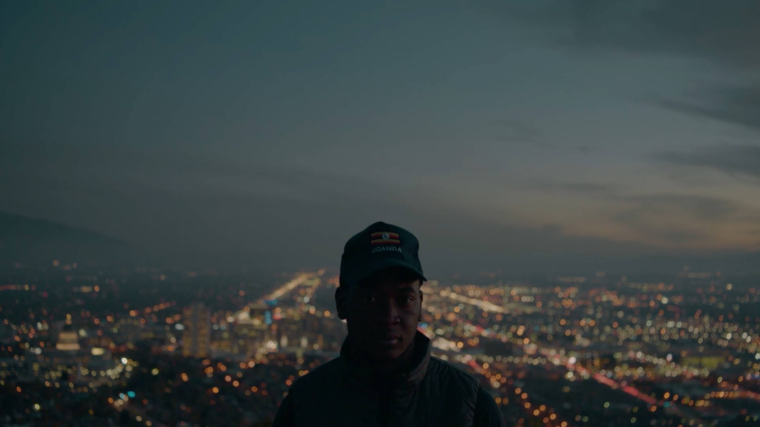 Cinematographer stands before city lights at dusk, wearing a hat with Uganda flag and text.